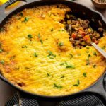A skillet of shepherd’s pie topped with golden mashed potatoes, partially served to show the meat and vegetable filling, with a spoon and garnished with chopped parsley.