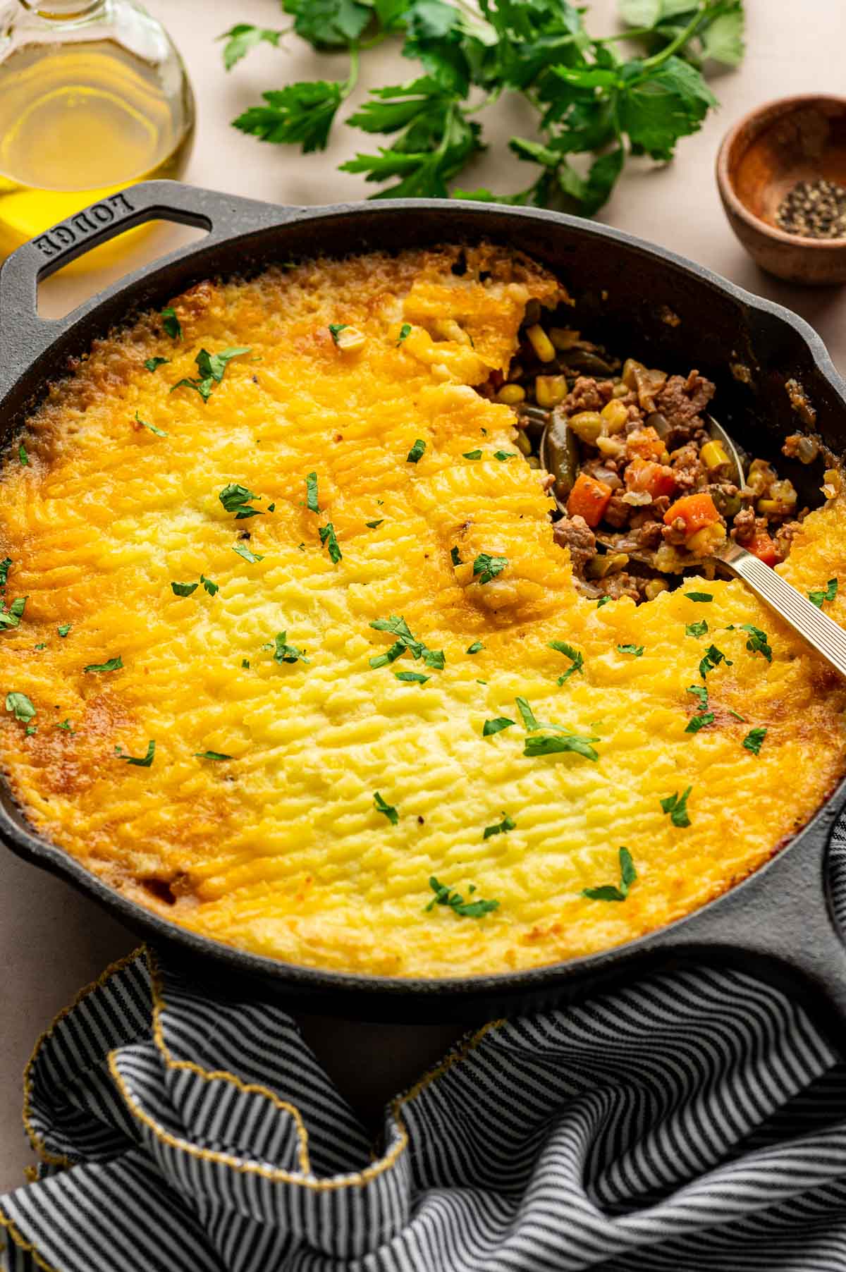 A skillet of shepherd’s pie topped with golden mashed potatoes, partially served to show the meat and vegetable filling, with a spoon and garnished with chopped parsley.