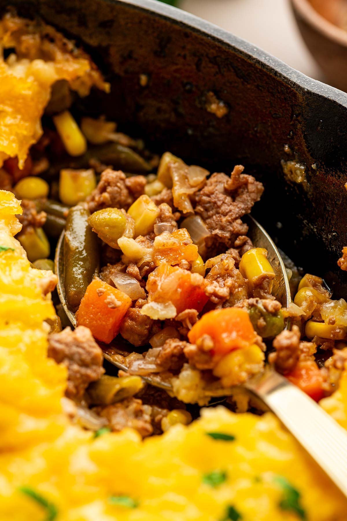 Close-up of a serving spoon scooping up classic shepherd's pie with ground lamb, mixed vegetables, and a mashed potato topping from a cast iron dish.