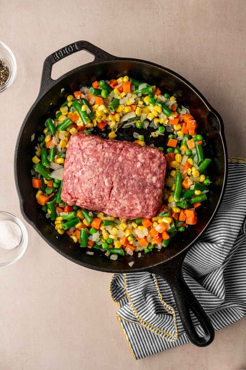 A cast iron skillet with mixed vegetables and a block of raw ground lamb in the center, placed on a light surface with a striped cloth nearby.