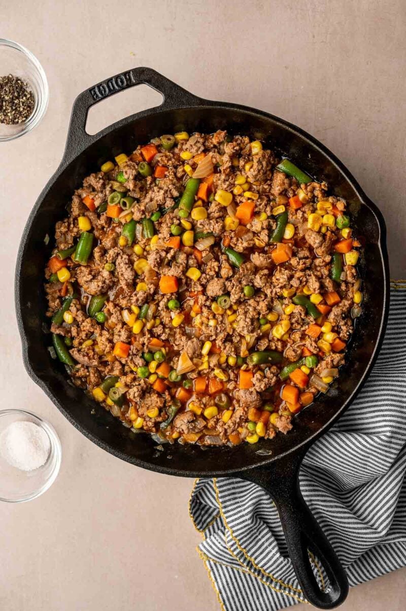 A cast iron skillet filled with cooked ground lamb, corn, green beans, peas, and carrots, set on a beige surface beside a striped cloth and small bowls of salt and pepper.