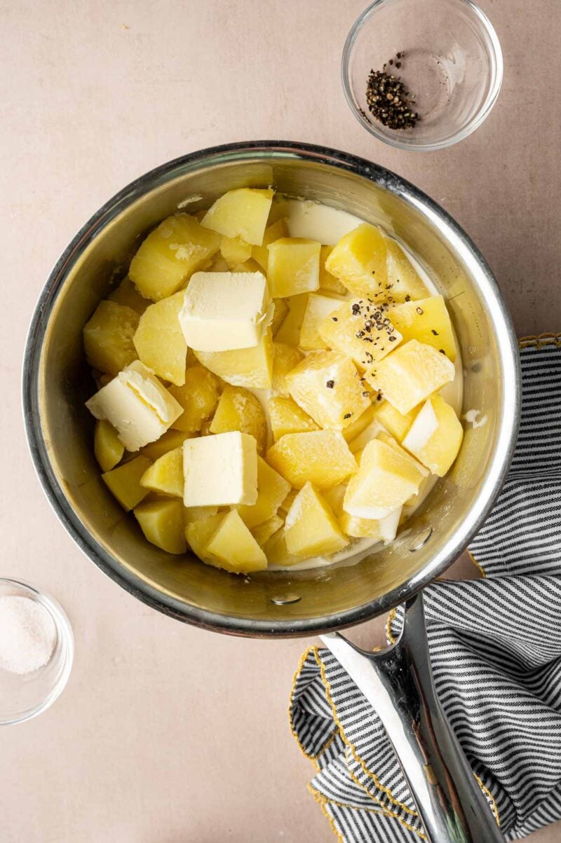 A pot filled with chopped cooked potatoes, butter, and black pepper sits on a counter near a striped towel, with small bowls of salt and pepper nearby.