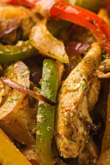 Close-up of cooked chicken strips with sliced red, yellow, and green bell peppers and onions, seasoned with herbs and spices.