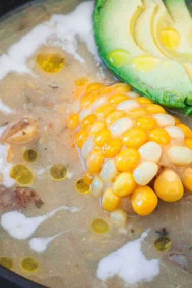 A bowl of soup with a piece of corn on the cob, avocado slices, and a drizzle of cream or oil on top.