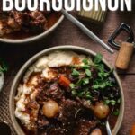 A bowl of beef bourguignon served over mashed potatoes, garnished with herbs, with a fork and a napkin on a wooden table.