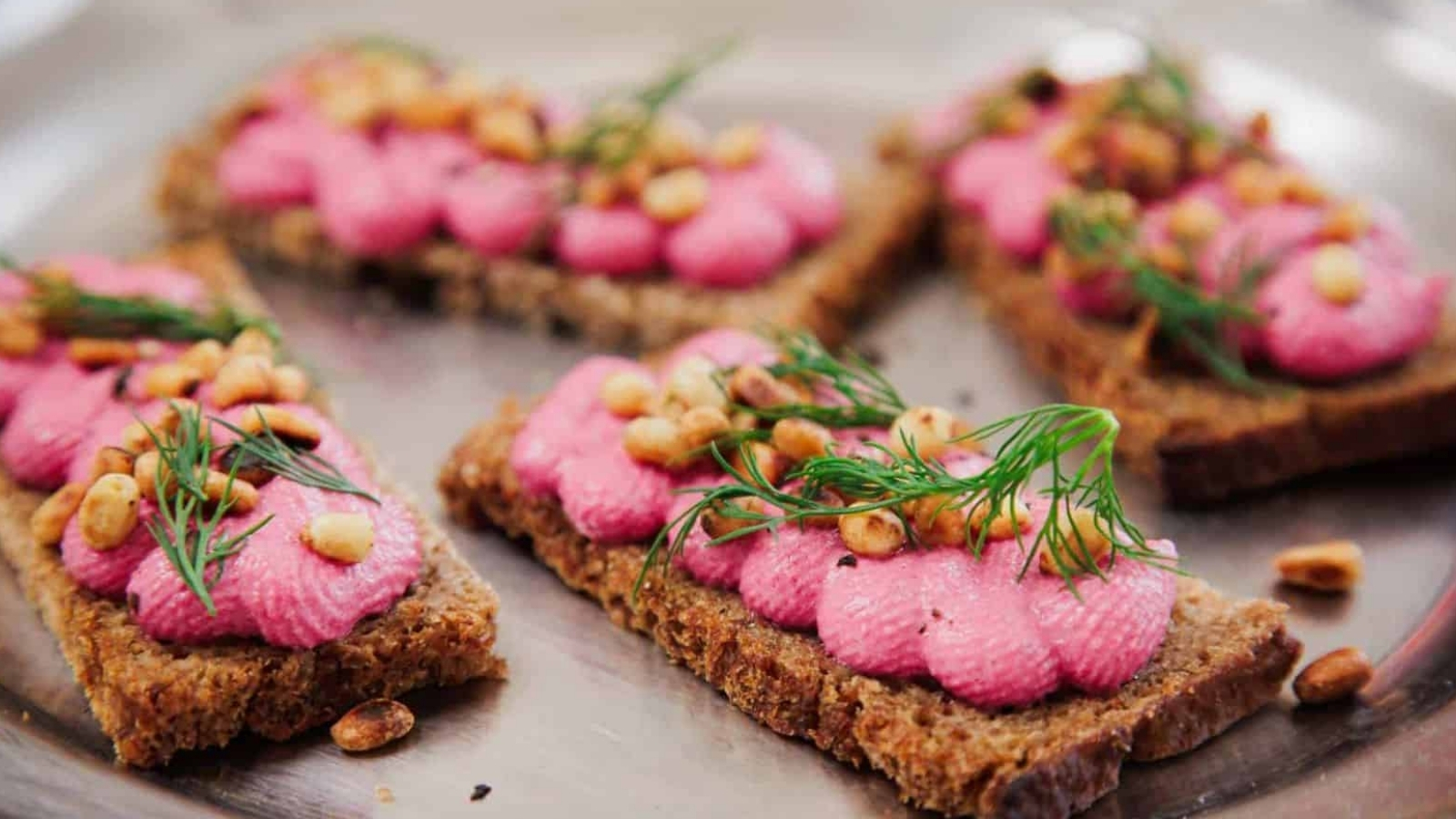 Four rectangular pieces of brown bread topped with pink spread, toasted pine nuts, and fresh dill are arranged on a metal tray.