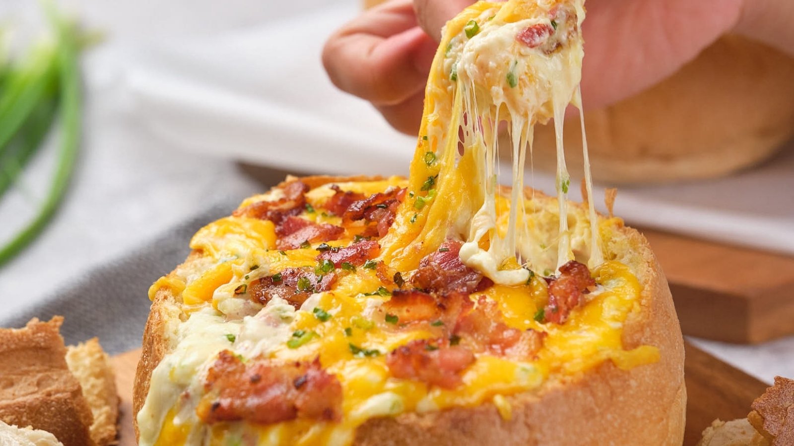 A person pulls a cheesy topping from a bread bowl filled with melted cheese and bacon bits.