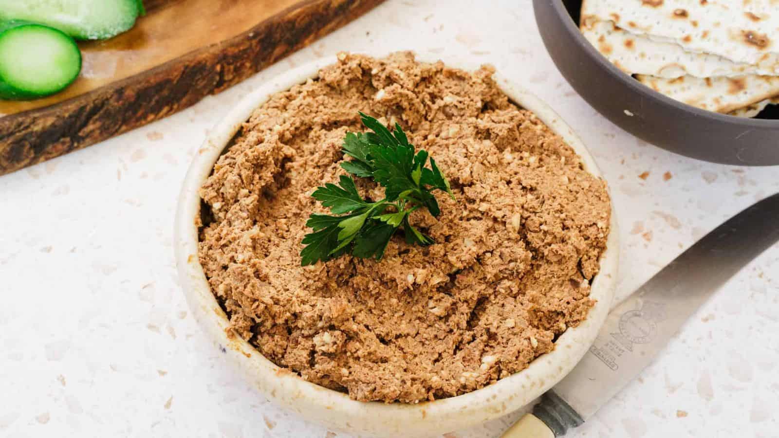 A bowl filled with a brown, chunky spread garnished with a sprig of parsley. A knife lies beside the bowl and slices of cucumber and crackers are in the background on a light-colored surface.