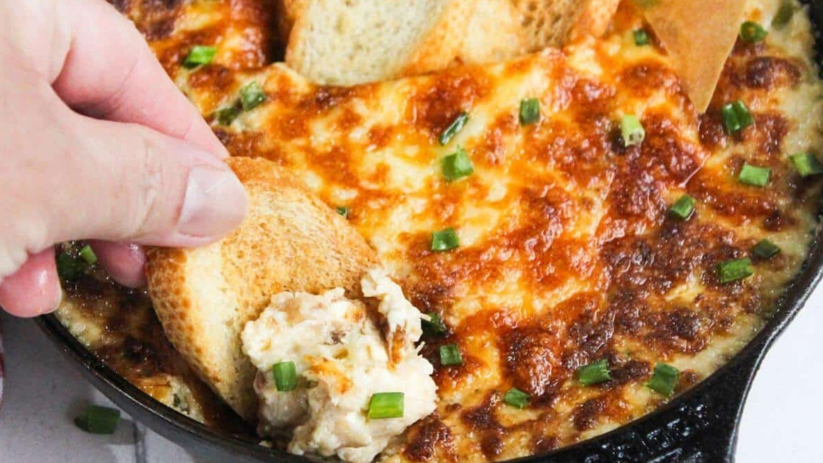 A person dipping a piece of bread into a skillet of cheesy dip.