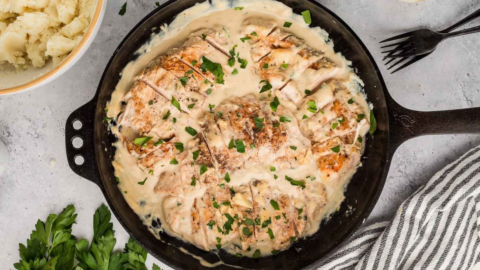 Sliced chicken breast in a creamy white sauce, garnished with chopped herbs, served in a cast iron skillet alongside mashed potatoes and utensils—a comforting classic among meat dinners.