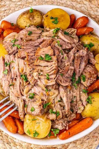 A white bowl filled with shredded pot roast, baby potatoes, and carrots, garnished with chopped parsley, on a woven placemat.