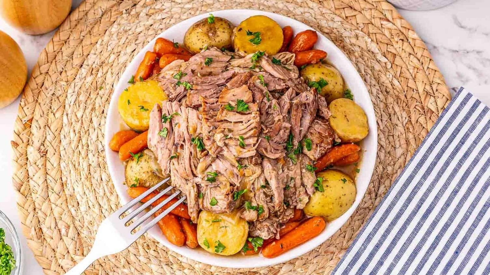 A bowl of shredded pot roast with baby potatoes and carrots, garnished with parsley, on a woven placemat.