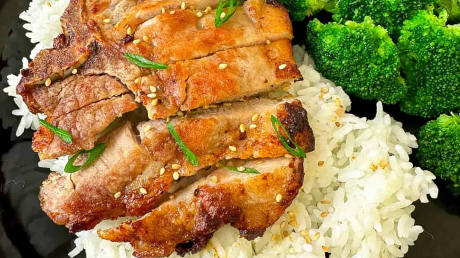 Grilled pork slices on white rice, garnished with sesame seeds and green herbs, served with broccoli florets.