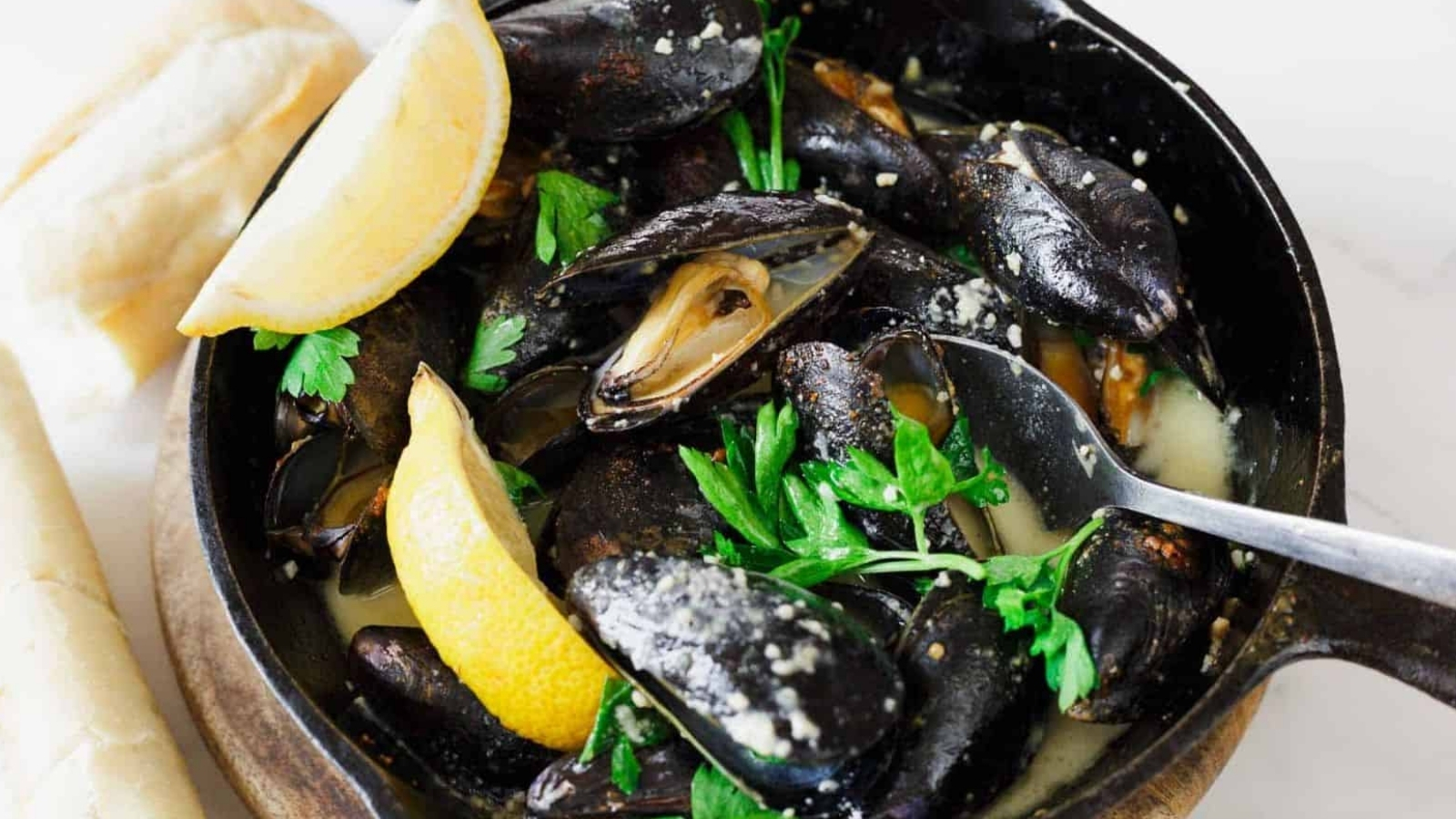 A skillet contains cooked mussels in a light sauce, garnished with parsley. A lemon wedge is placed inside, and a spoon rests in the dish. Partially visible bread rolls are on the side.