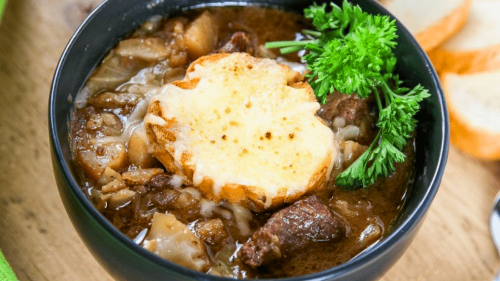 A bowl of neef stew with bread topped with melted cheese and a sprig of parsley, placed next to sliced bread.