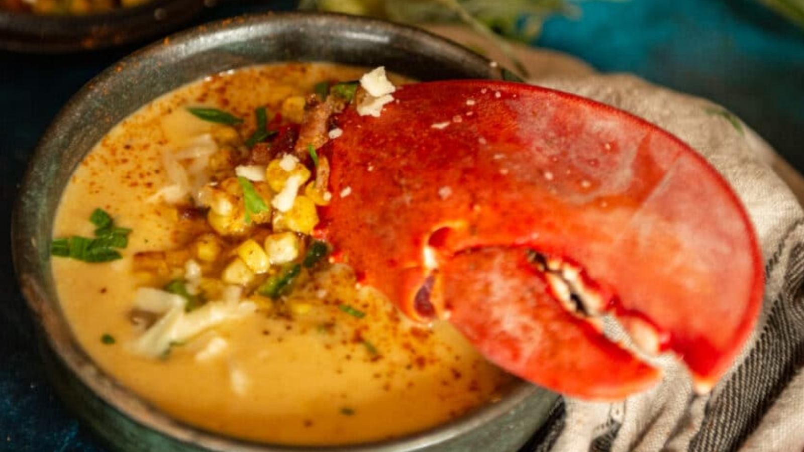 A bowl of creamy soup garnished with corn, herbs, and cheese, topped with a large whole lobster claw.