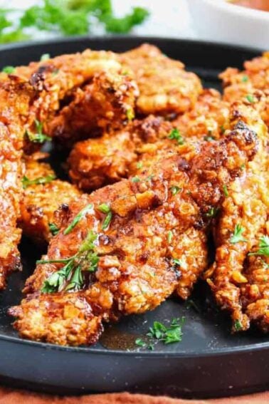 A black plate filled with several crispy, golden-brown chicken tenders garnished with chopped parsley, set on a brown cloth with sauce in the background.