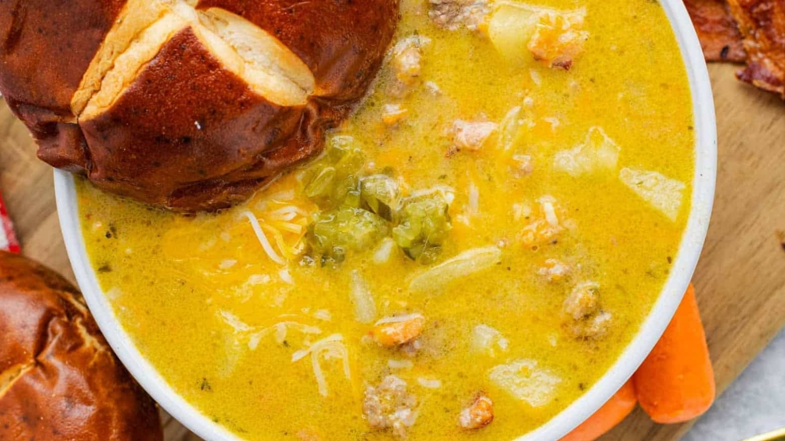 A bowl of cheesy soup with ground meat, potatoes, and vegetables, served with a pretzel bun.