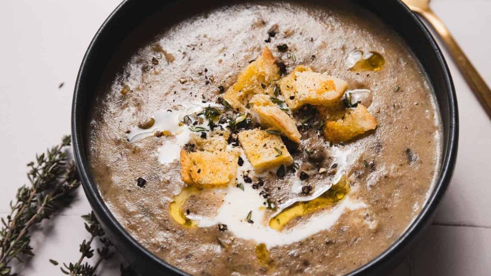 A bowl of creamy mushroom soup garnished with croutons, a drizzle of olive oil, and fresh herbs. A golden spoon lies nearby, and sprigs of thyme are placed on the table beside the bowl.