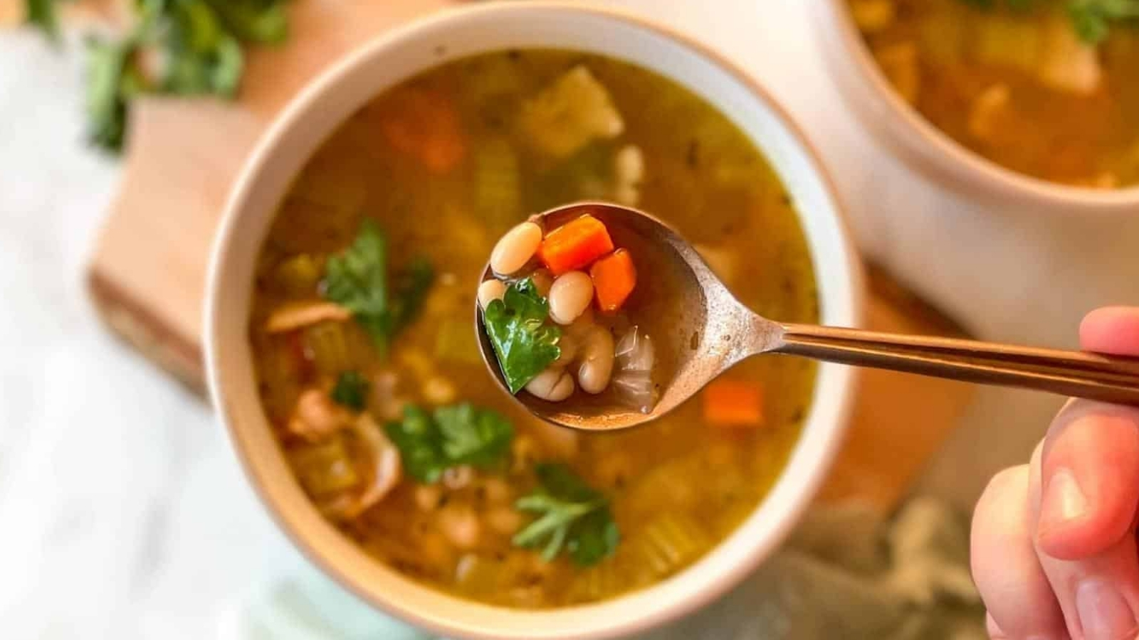 A hand holds a spoonful of vegetable soup with beans, carrots, celery, and herbs above a bowl of the same soup.