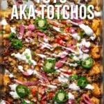 A sheet pan of loaded tater tots topped with ground meat, melted cheese, jalapeños, tomatoes, onions, and a drizzle of white sauce.