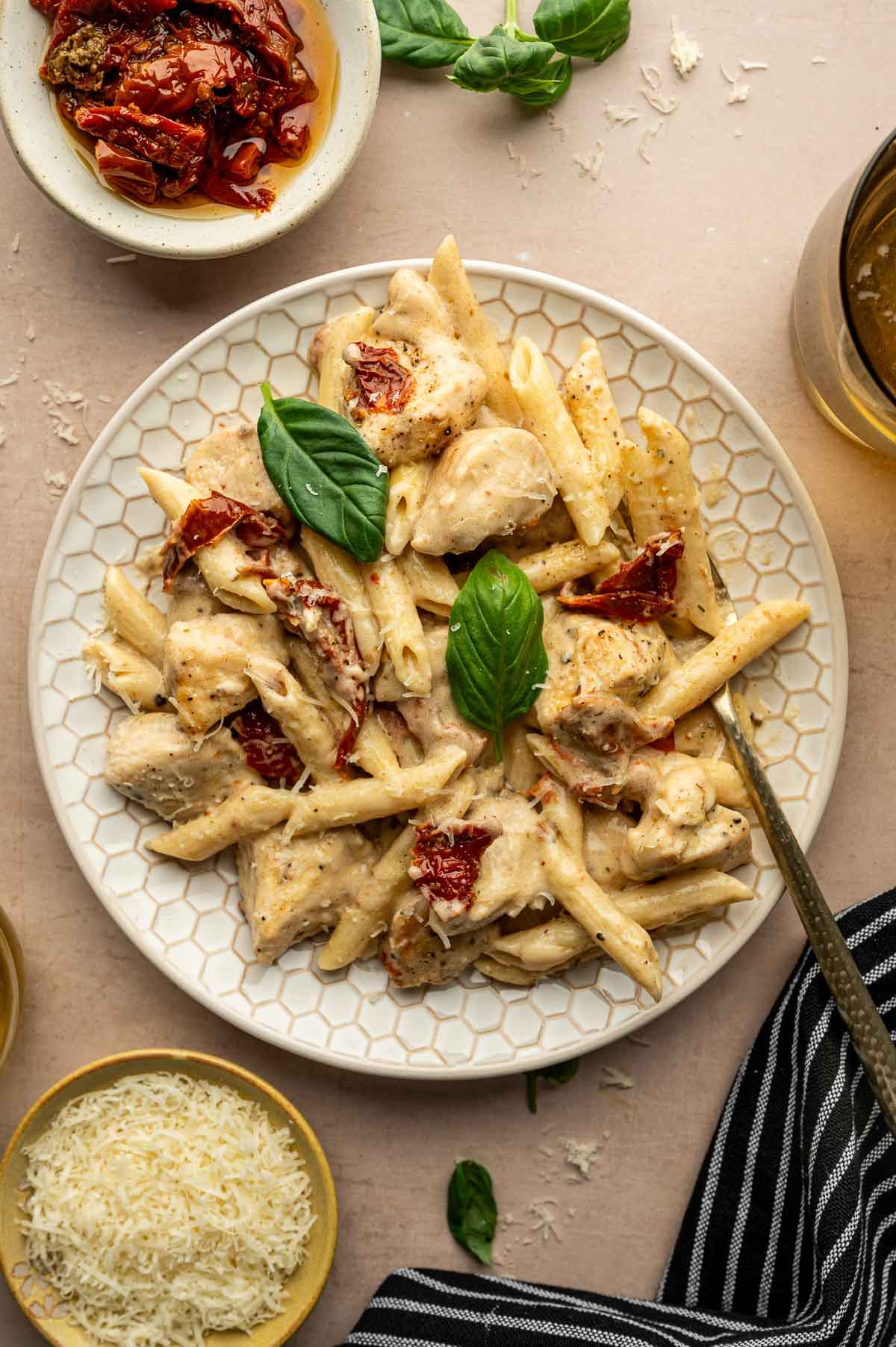A plate of marry me pasta with pieces of chicken, sun-dried tomatoes, creamy sauce, and a basil garnish, surrounded by grated cheese, sun-dried tomatoes, and a drink.