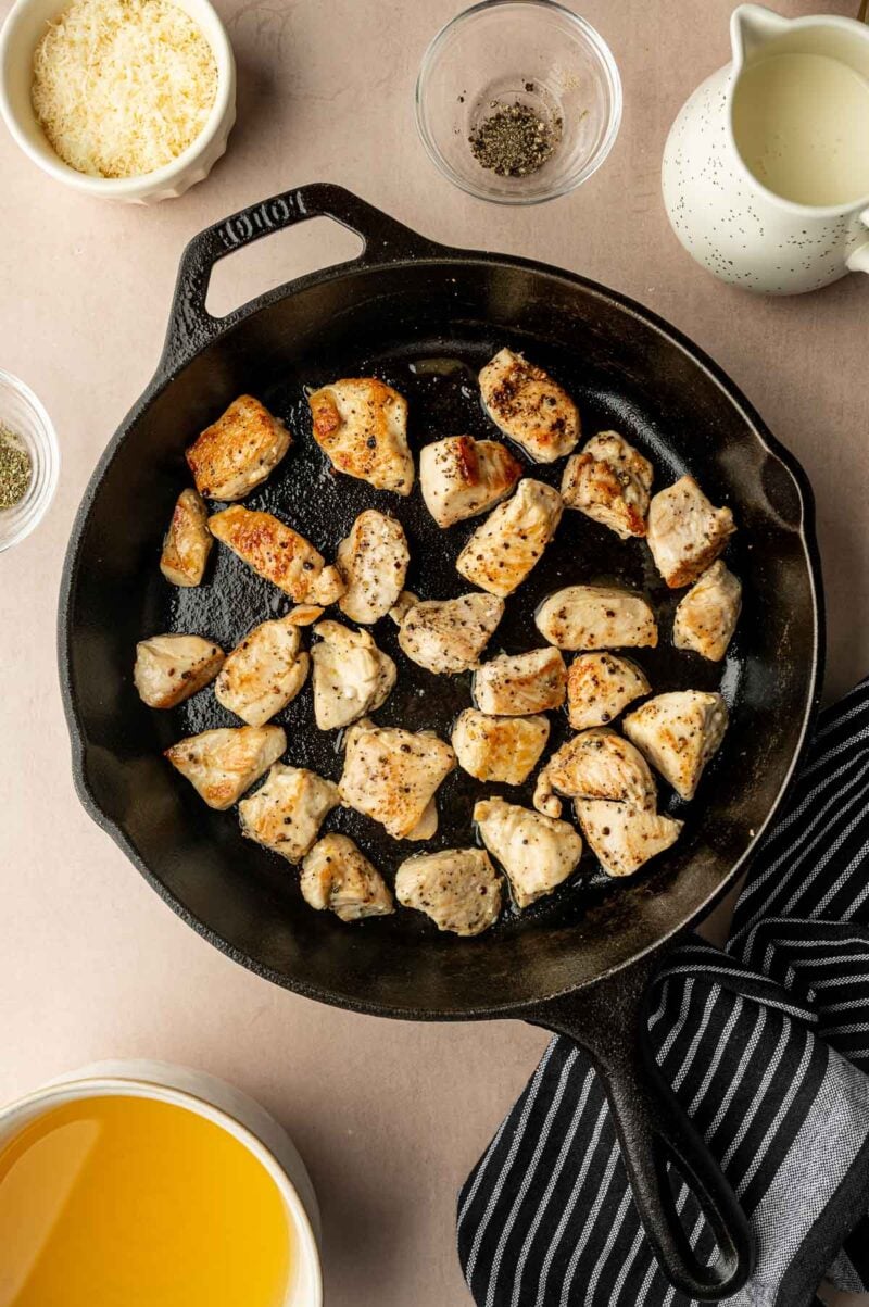 Chunks of cooked chicken breast in a black cast iron skillet, surrounded by bowls of shredded cheese, black pepper, broth, and a striped cloth napkin.