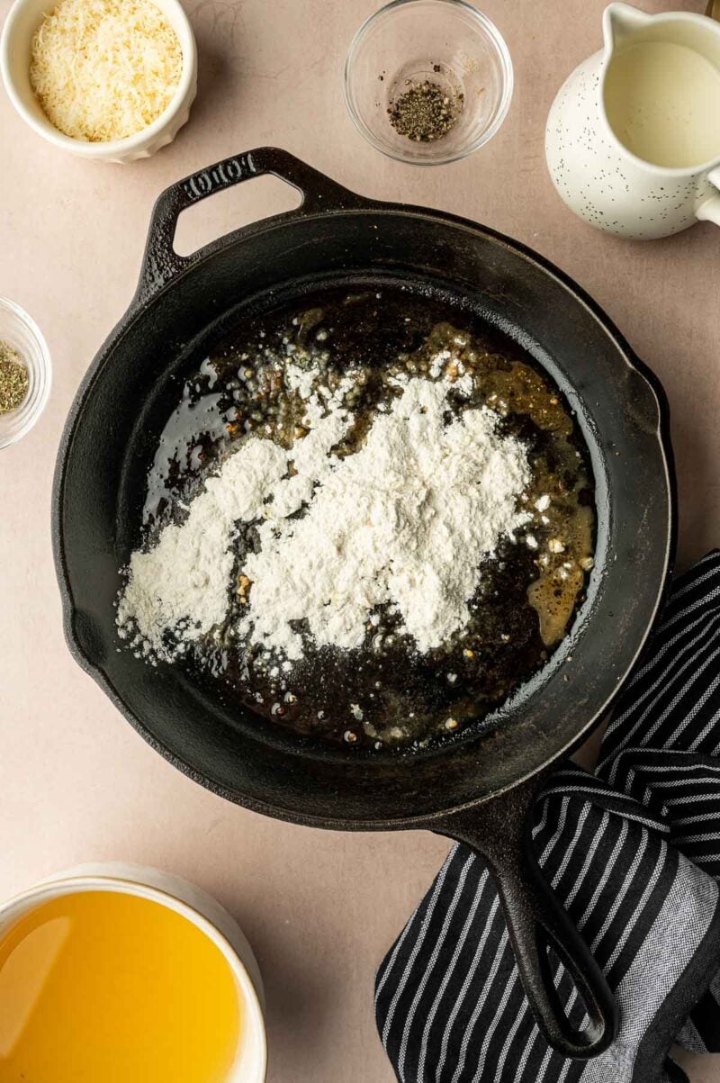 A cast iron skillet with melted butter and flour, surrounded by bowls of broth, grated cheese, pepper, and a pitcher of milk on a countertop.