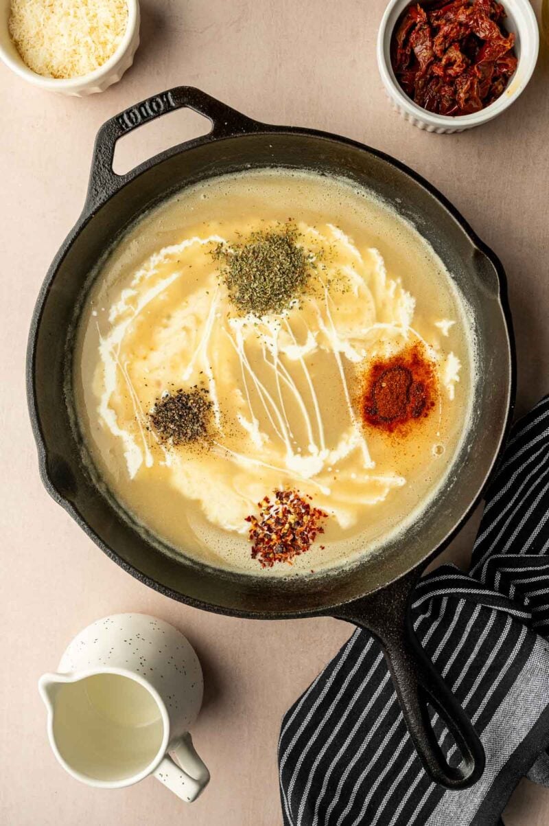 A cast iron skillet contains a creamy sauce with dried herbs, chili flakes, paprika, and black pepper. Surrounding the skillet are bowls of sun-dried tomatoes, grated cheese, and a small pitcher of cream.