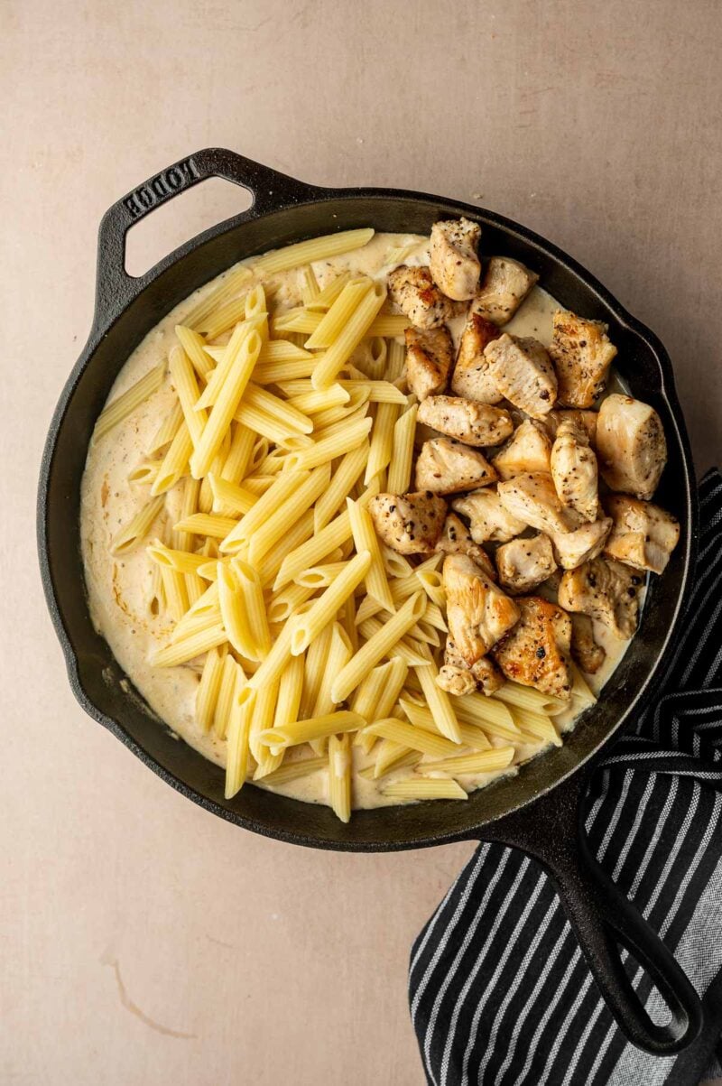 A cast iron skillet with cooked penne pasta, grilled chicken pieces, and creamy sauce, placed on a light surface next to a striped napkin.
