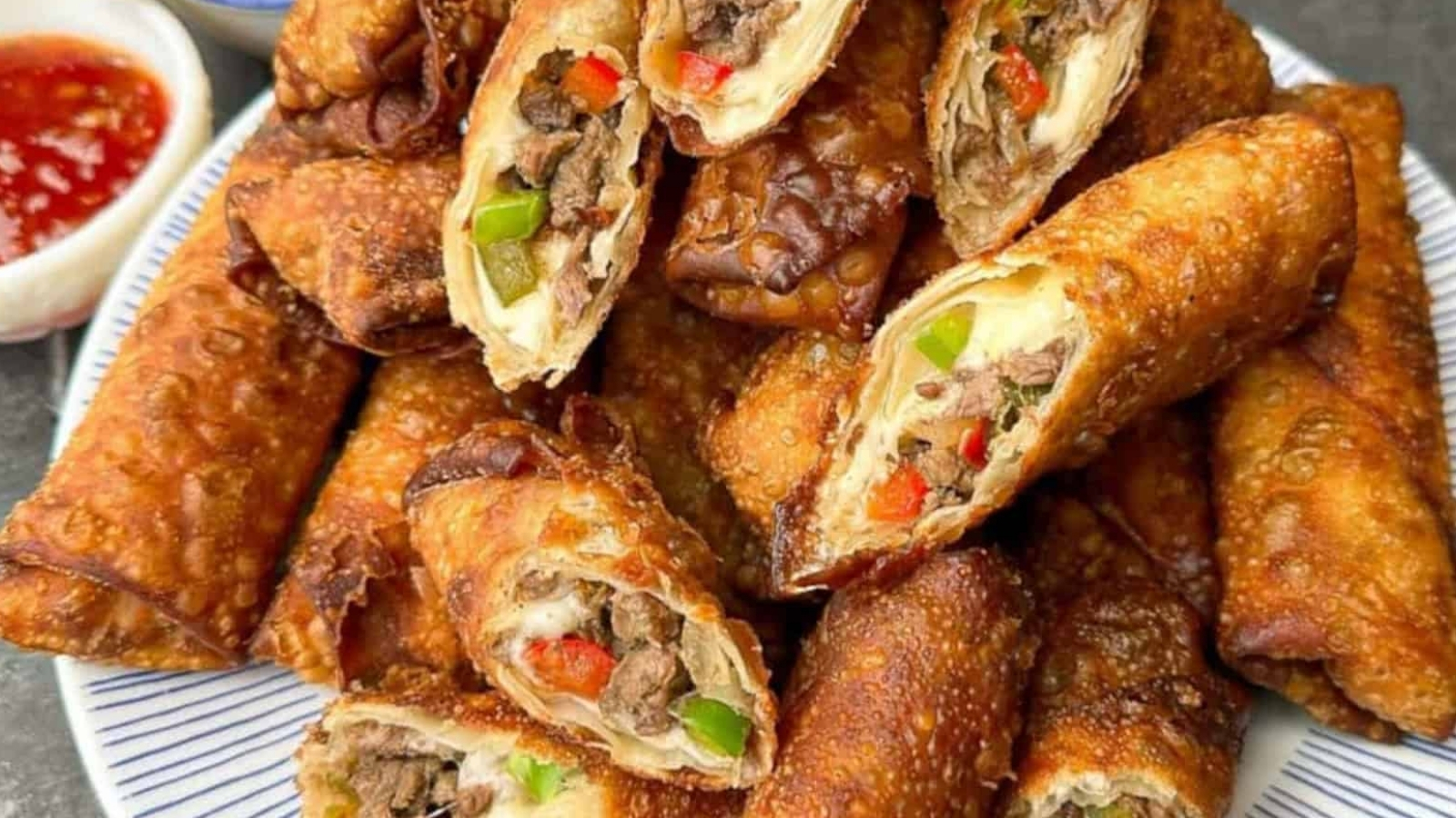 A plate of crispy egg rolls filled with beef, green and red peppers. A small dish of dipping sauce is visible on the side.
