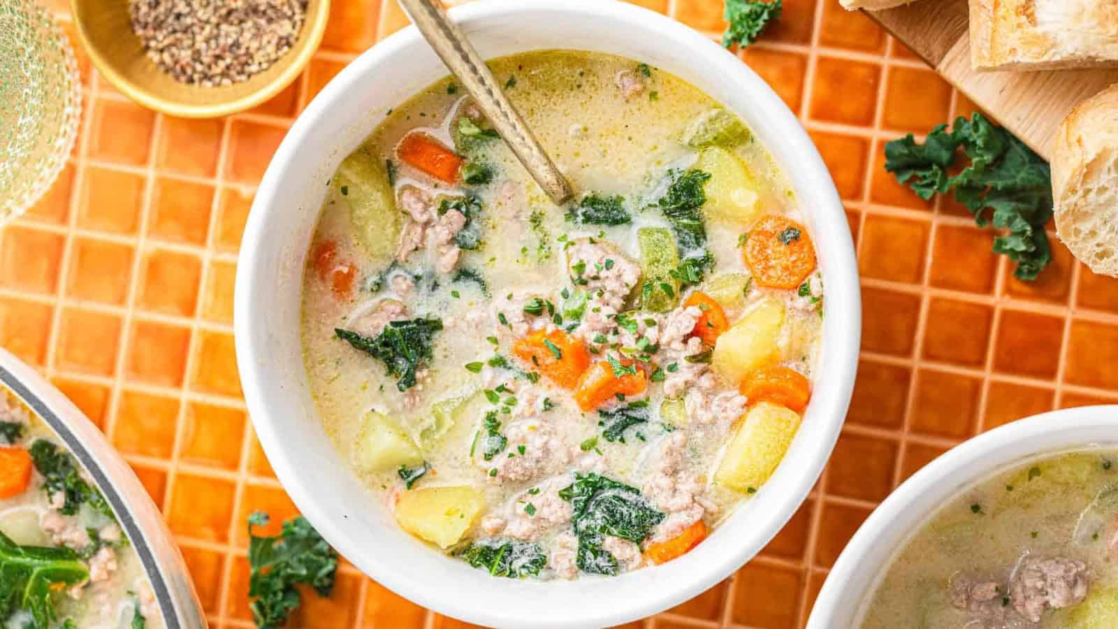 A bowl of creamy soup with sausage, kale, carrots, and potatoes, with a spoon inside, on an orange tiled surface.