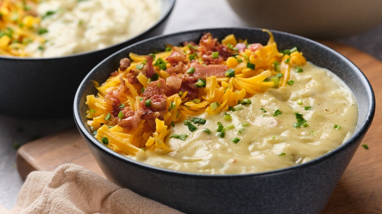 A bowl of creamy potato soup topped with shredded cheddar cheese, bacon bits, and chopped chives, with another bowl in the background.