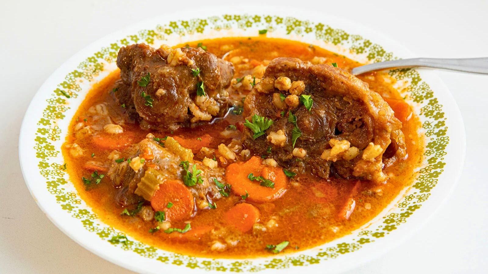 A bowl of oxtail stew with carrots, celery, rice, and parsley in a rich, reddish broth, served on a patterned plate with a spoon.