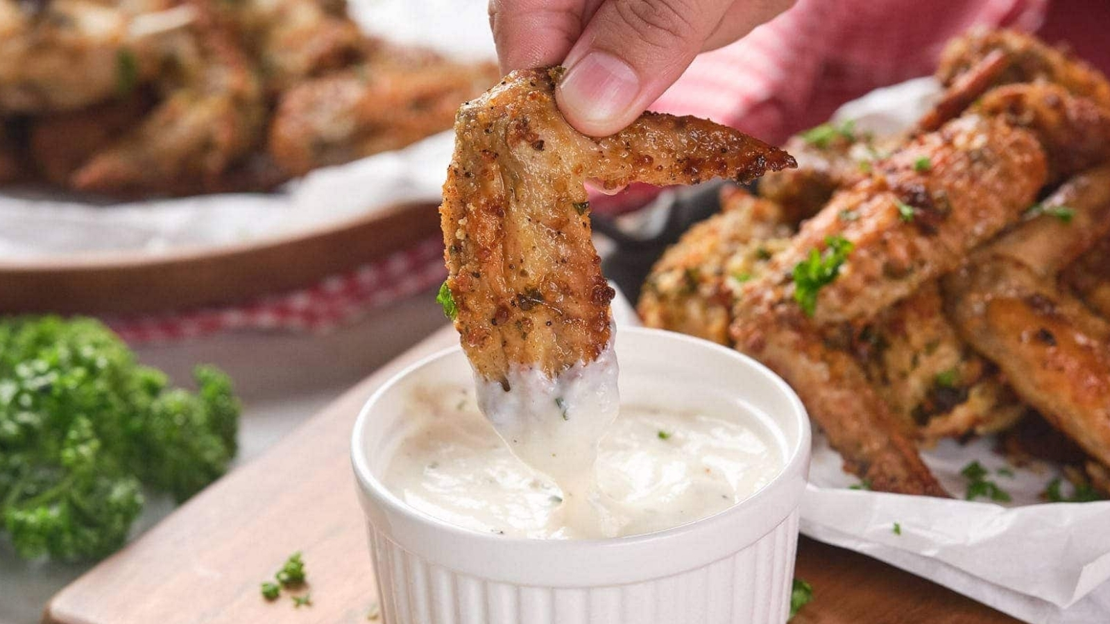 A hand dips a parmesan chicken wing into a small ramekin filled with white dipping sauce.