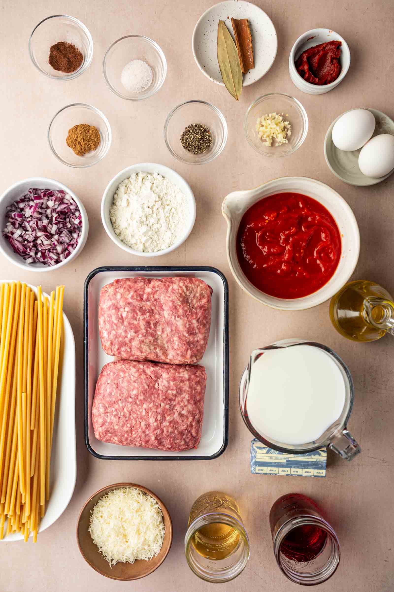A top-down view of ingredients for a pasta dish, including ground lamb, pasta, eggs, tomato sauce, milk, cheese, olive oil, spices, and seasonings arranged neatly on a countertop.