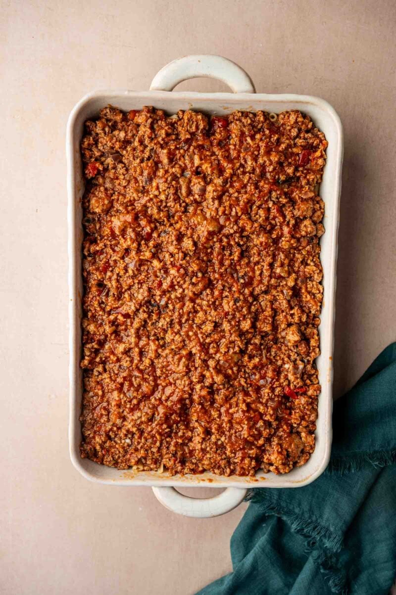 A rectangular white casserole dish filled with a ground lamb mixture, placed on a light surface next to a dark green cloth.
