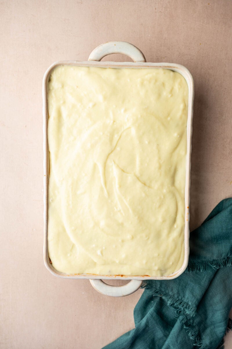 A rectangular baking dish filled with a smooth, creamy béchamel mixture sits on a light surface next to a teal cloth.