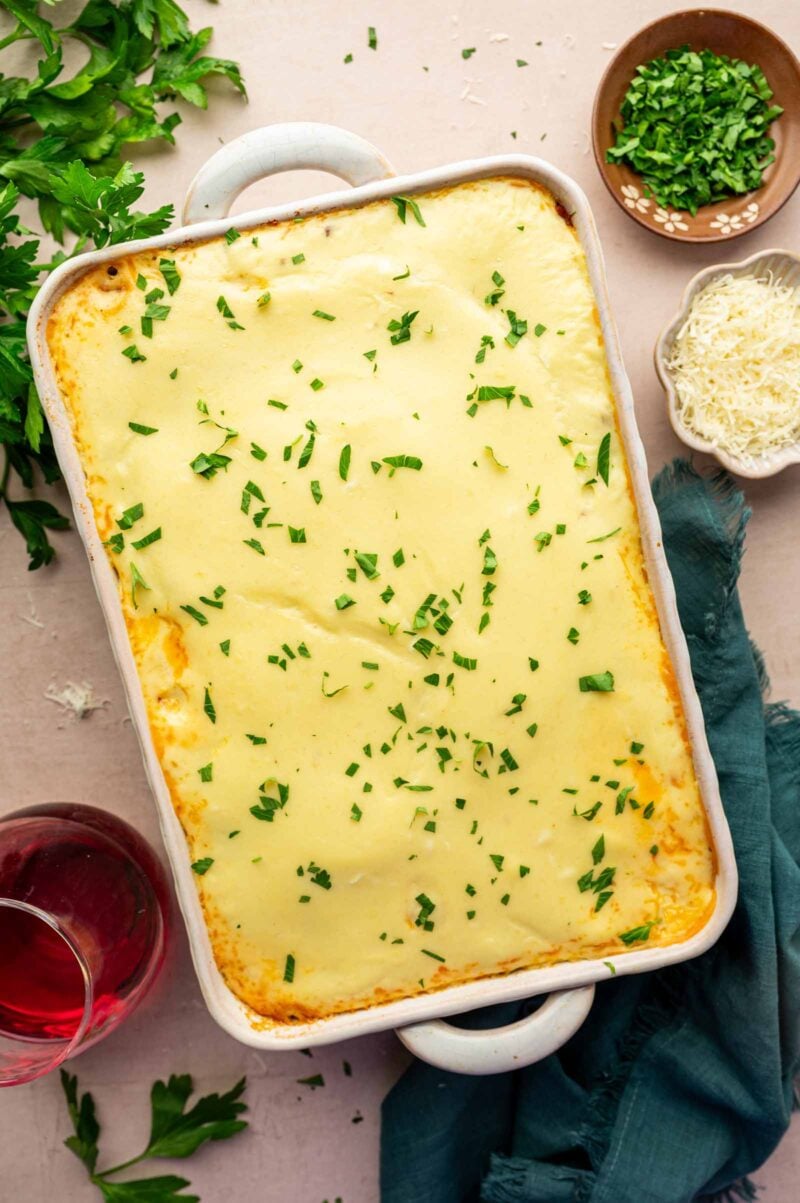 A rectangular casserole dish filled with a baked, creamy pastitsio topped with béchamel chopped green herbs. Shredded cheese, chopped herbs, and a glass of red liquid are nearby.