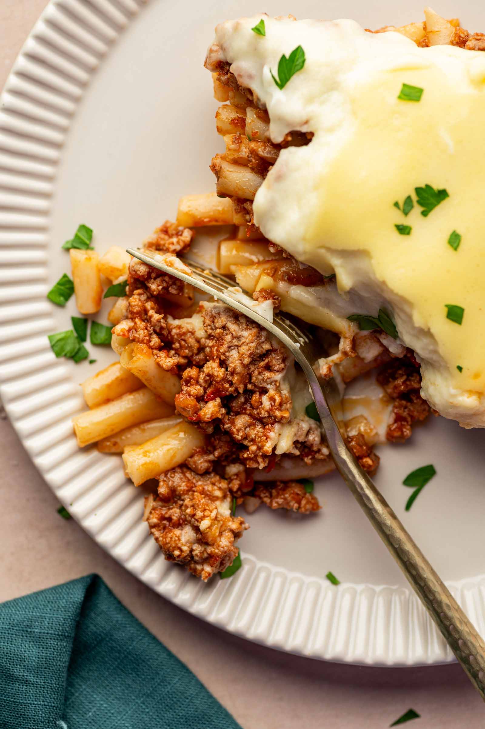 A close-up of a plated baked pasta dish topped with meat sauce and creamy white sauce, garnished with chopped herbs, with a fork cutting through the serving.