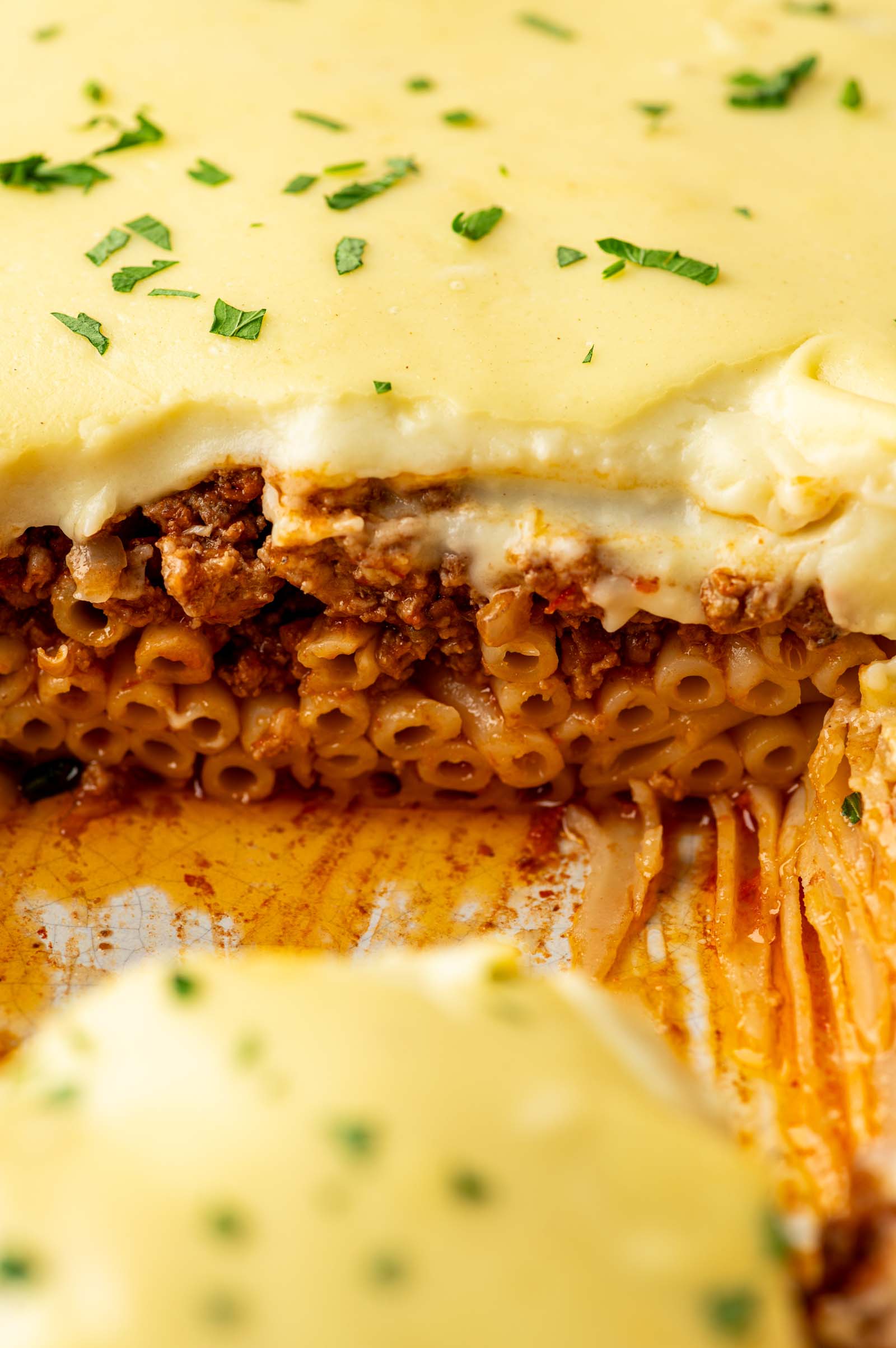 A close-up of a sliced baked pastitsio dish topped with creamy sauce, ground lamb, and garnished with chopped parsley.