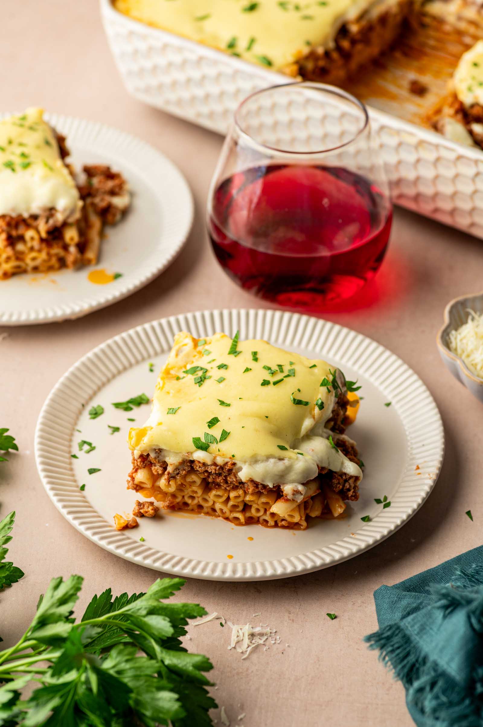 A square serving of baked pastitsio with meat sauce and béchamel on a white plate, alongside a glass of red wine and garnished with chopped parsley.