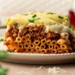 A slice of baked pastitsio with layered noodle pasta, ground lamb sauce, and béchamel sauce, garnished with chopped parsley on a white plate.