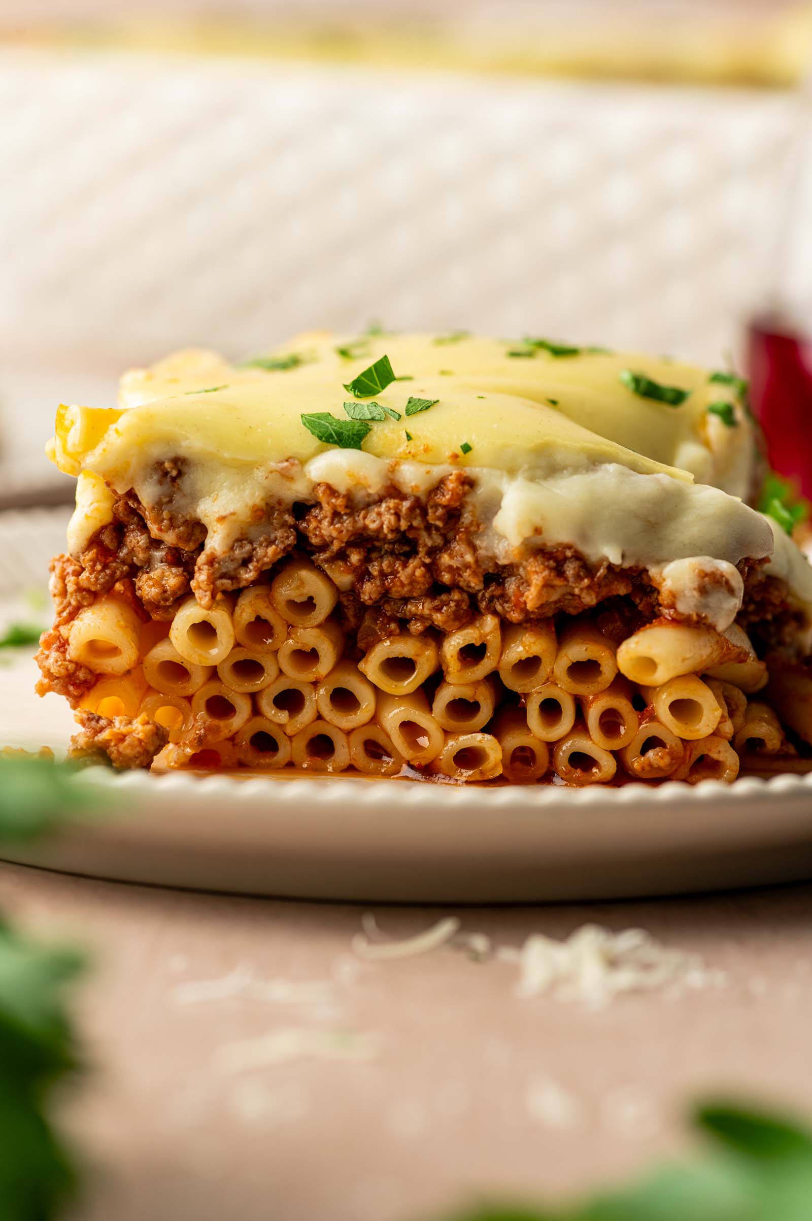 A slice of baked pastitsio with layered noodle pasta, ground lamb sauce, and béchamel sauce, garnished with chopped parsley on a white plate.