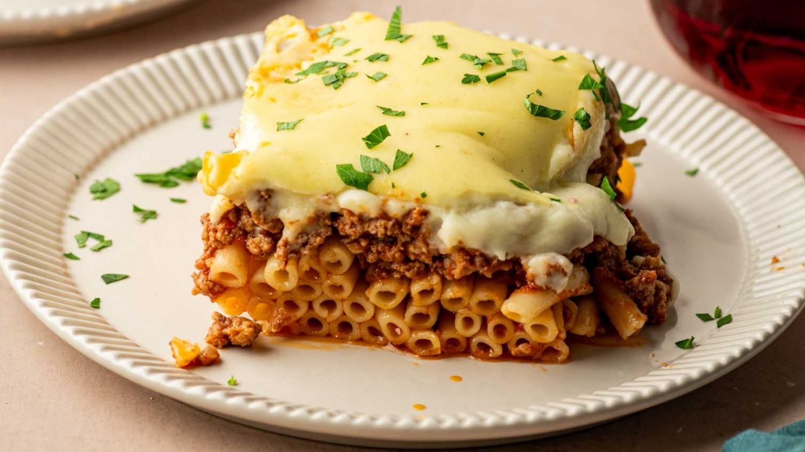 A square slice of pastitsio with ground meat, pasta, and creamy béchamel sauce, garnished with chopped parsley, served on a white plate.