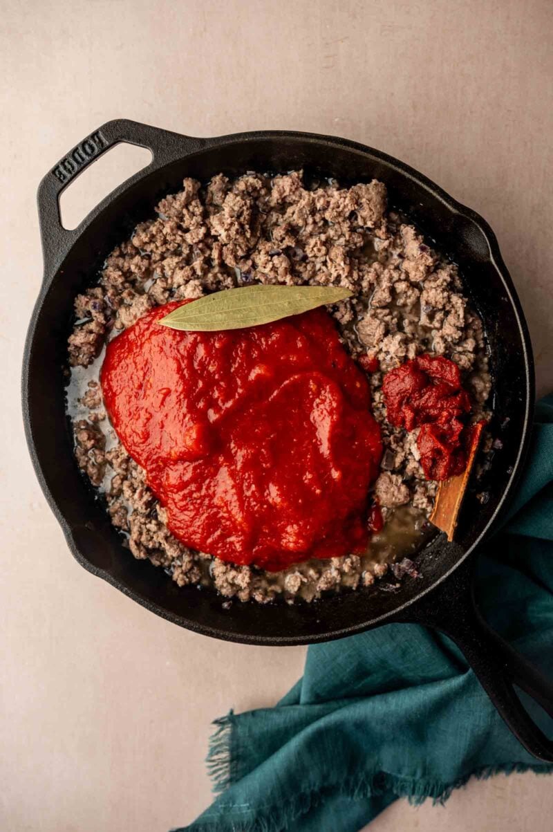 A cast iron skillet with cooked ground lamb, tomato sauce, tomato paste, and a bay leaf, placed on a light surface with a green cloth nearby.