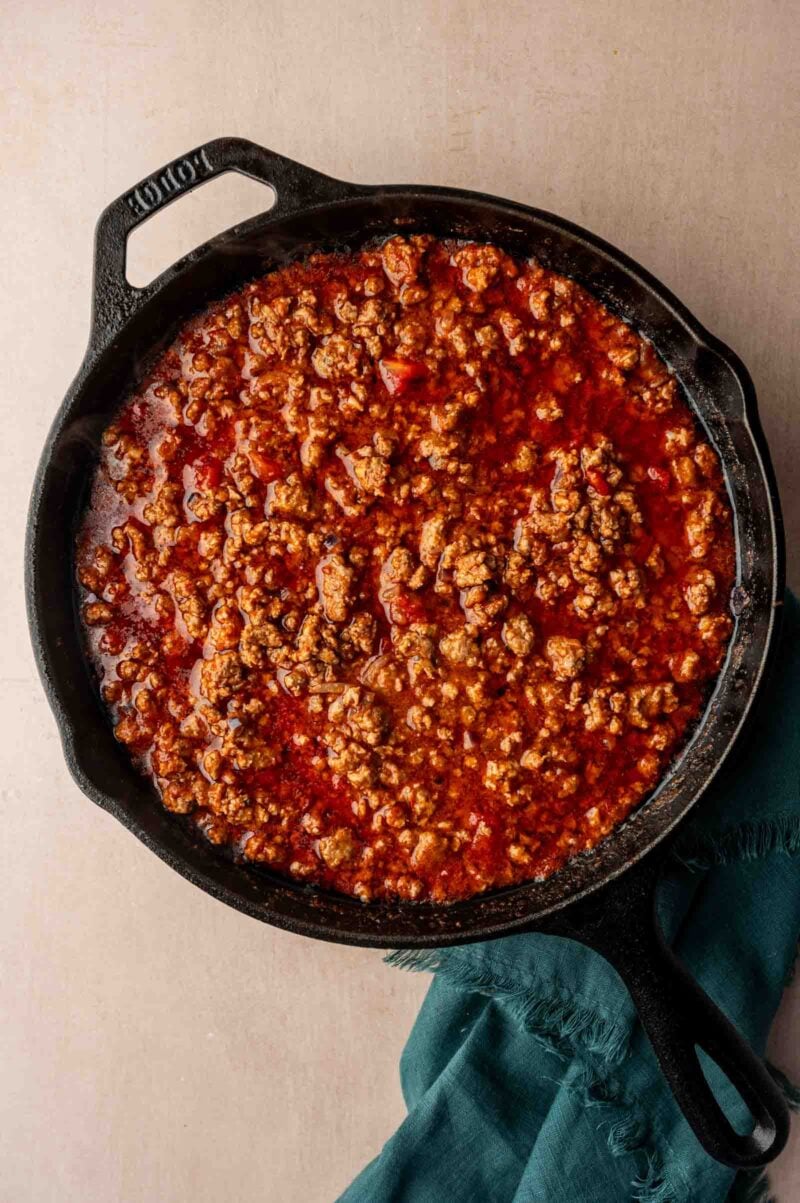 Ground lamb cooked in tomato sauce in a black cast iron skillet, placed on a light surface with a dark green cloth partially under the pan.