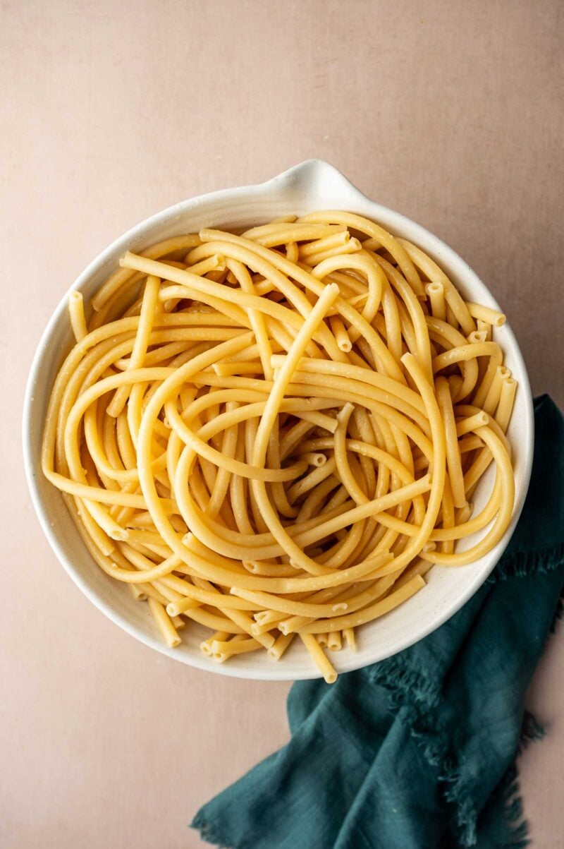 A white bowl filled with cooked bucatini pasta sits on a light surface next to a dark green cloth napkin.