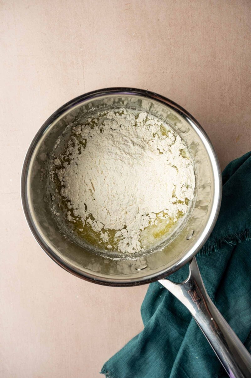 A metal saucepan containing melted butter and flour sits on a light surface next to a green cloth.