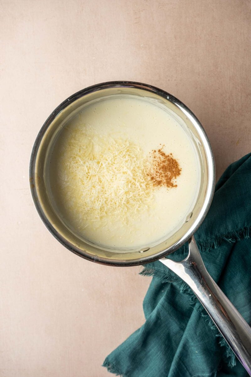 A saucepan filled with creamy white sauce topped with grated cheese and a sprinkle of ground nutmeg, resting on a beige surface next to a green cloth.
